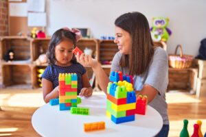 Beautiful BCBA and toddler girl playing with construction blocks building tower at kindergarten