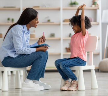 5 Steps on How to Become a BCBA Excited cute little african american girl at child psychologists office, sitting on chair in front of attractive female therapist, raising hands up, smiling, feeling safe. Child psychotherapy concept