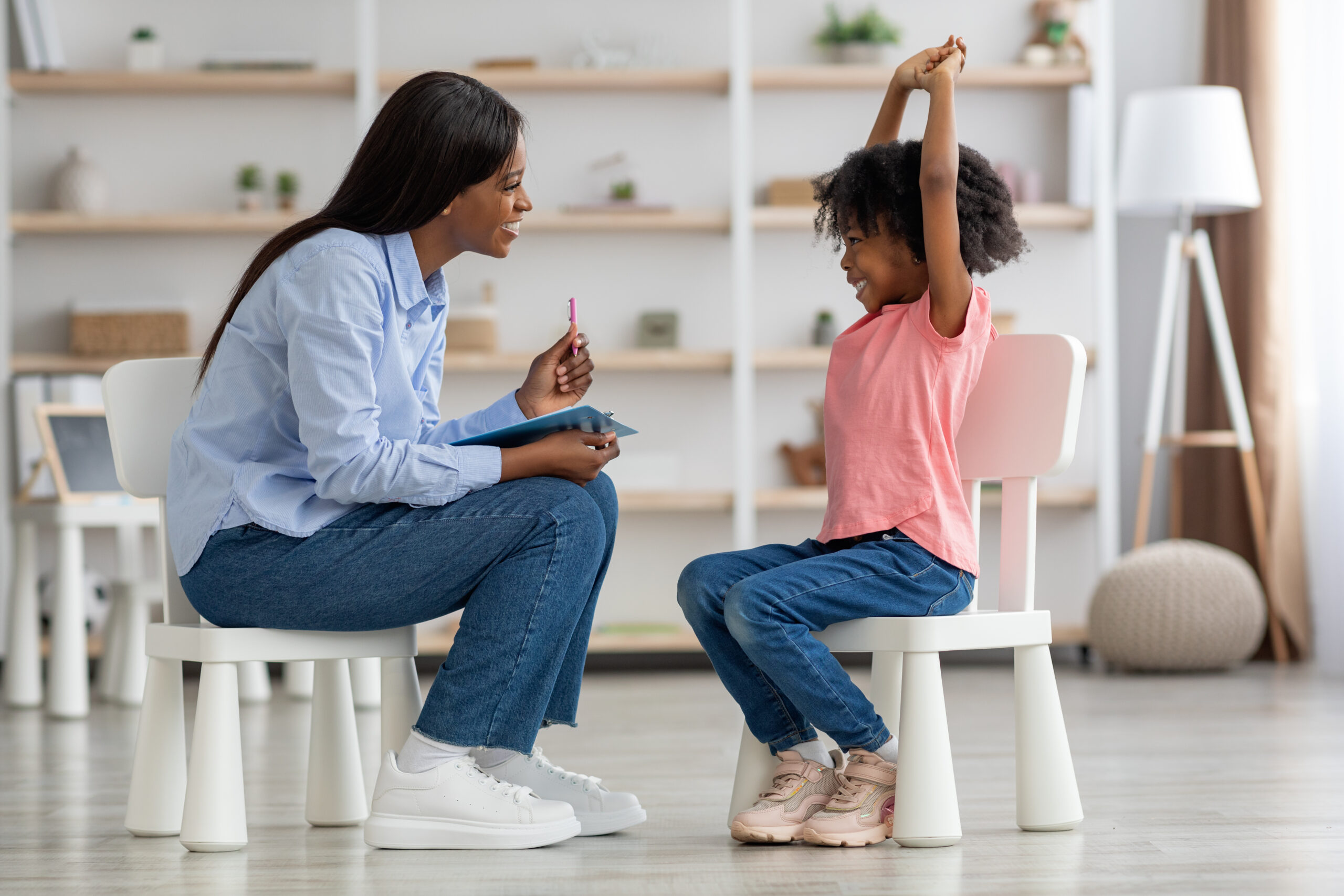 5 Steps on How to Become a BCBA Excited cute little african american girl at child psychologists office, sitting on chair in front of attractive female therapist, raising hands up, smiling, feeling safe. Child psychotherapy concept