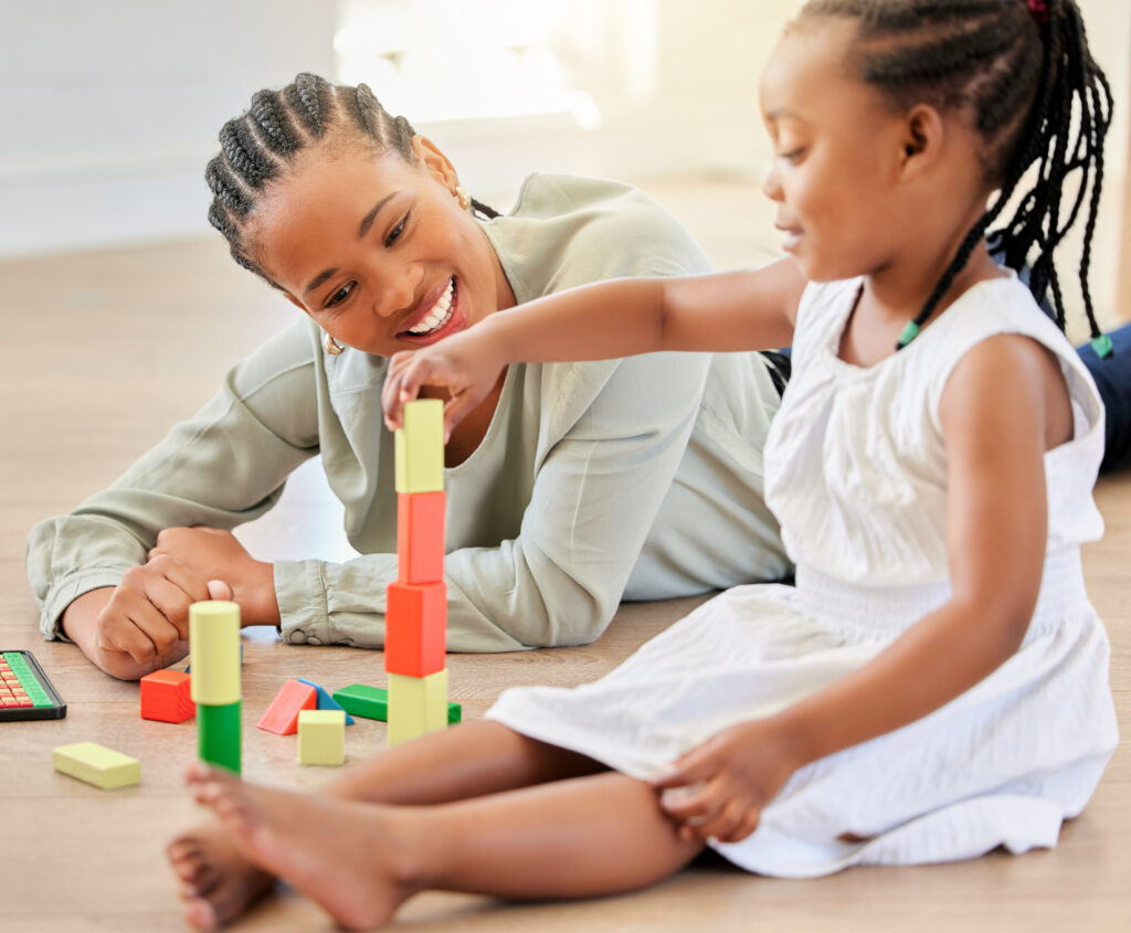 African american girl playing with building blocks while sitting with mother. Little girl building tower with wooden blocks. Smiling woman lying on the floor and playing with child.