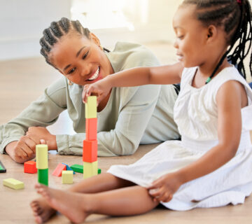 African American girl BCBA playing with building blocks while sitting with mother doing ABA therapy. Little girl building tower with wooden blocks. Smiling BCBA woman lying on the floor and playing with child.