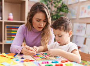 Behavior Technician Woman and Autistic boy playing with maths puzzle game sitting on table at kindergarten